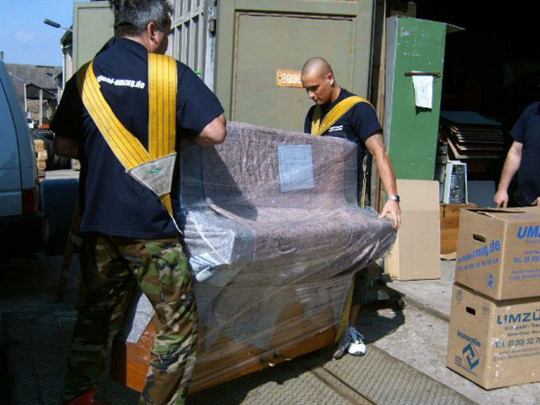 Mitarbeiter von Kranepuhl tragen ein in Folie verpacktes Klavier mit Tragegurten aus einem Gebäude
